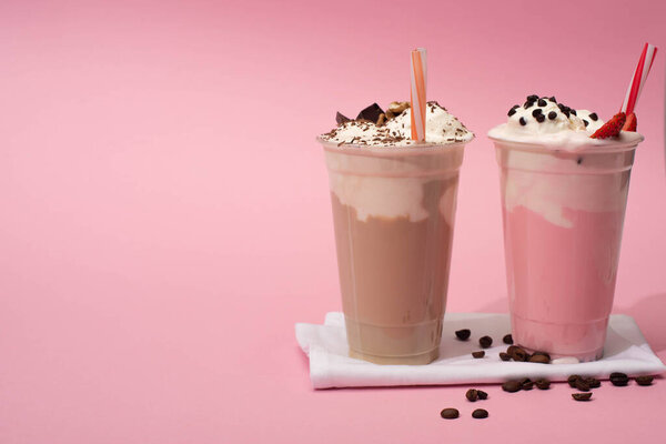 Disposable cups of chocolate and strawberry milkshakes with drinking straws and coffee grains on napkins on pink