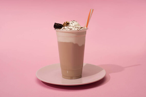 Disposable cup of chocolate milkshake with drinking straw on plate on pink background