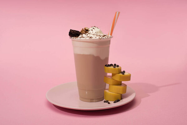Disposable cup of milkshake with drinking straw, cut banana and chocolate chips on plate on pink background