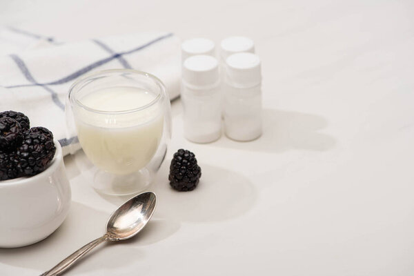 High angle view of teaspoon, glass of yogurt and sugar bowl with blackberries near containers with starter cultures on white