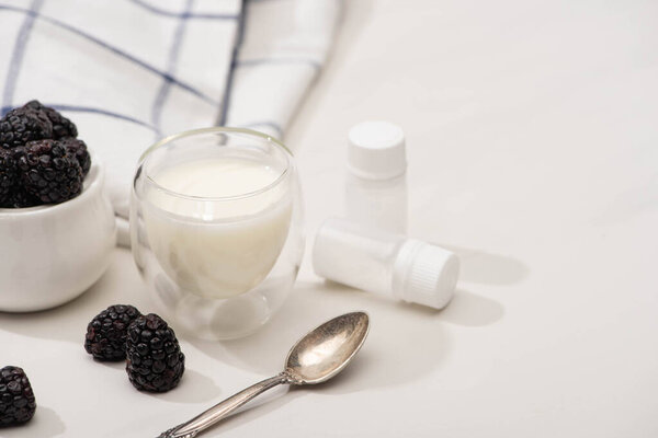 Selective focus of glass of yogurt, teaspoon, containers with starter cultures and sugar bowl with blackberries on white