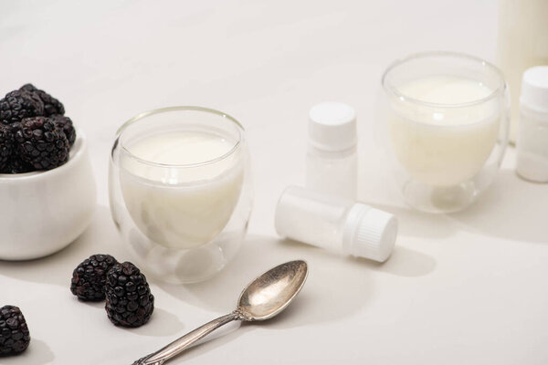 High angle view of glasses of yogurt, teaspoon, containers with starter cultures and sugar bowl with blackberries on white