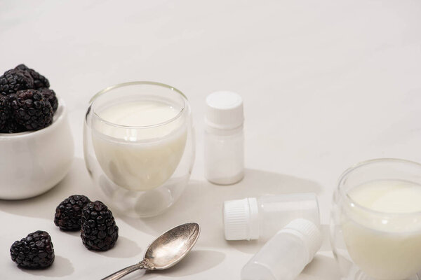High angle view of glasses of yogurt, teaspoon, containers with starter cultures near sugar bowl with blackberries on white