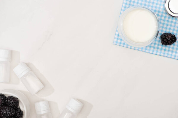 Top view of glass of yogurt on cloth, containers with starter cultures and blackberries on white background