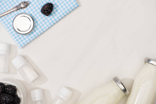 Top view of plaid cloth with lid and blackberry near containers with starter cultures and bottles of homemade yogurt on white