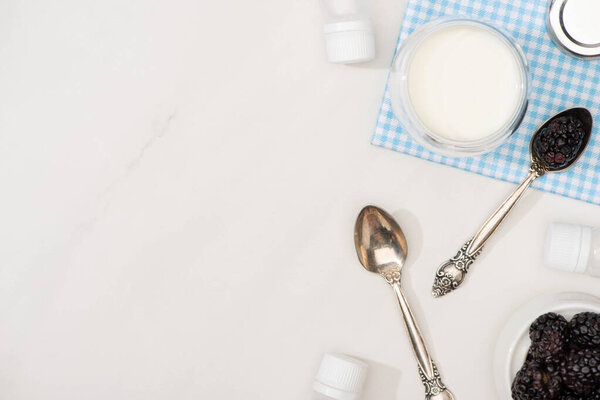 Top view of glass of yogurt on cloth, teaspoons, blackberries and containers with starter cultures on white