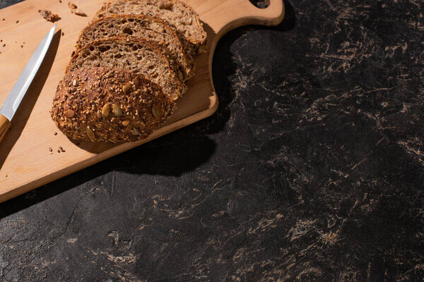 top view of cut whole grain bread on cutting board near knife on stone black surface