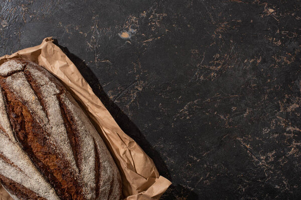 top view of organic brown bread loaf on paper on stone black surface