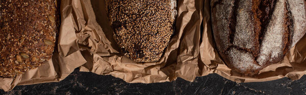 top view of fresh baked whole grain bread loaves in paper on stone black surface, panoramic shot