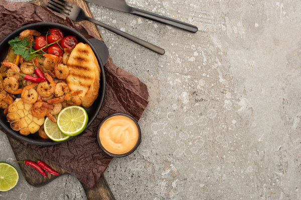 top view of fried shrimps with grilled toasts, vegetables and lime served on board near cutlery and sauce on grey concrete background