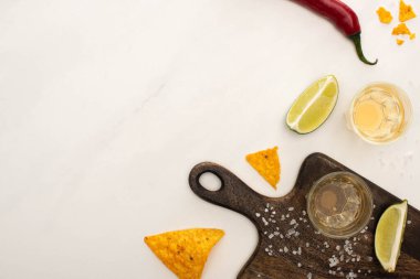 Beyaz mermer yüzey üzerinde ahşap kesim tahtasının yanında limonlu altın tekila, kırmızı biber, tuz ve nachos manzarası.