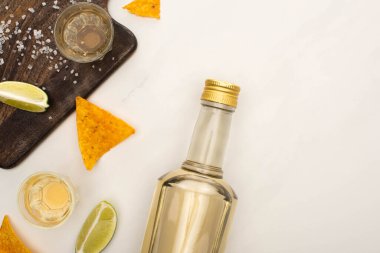 Beyaz mermer yüzeyde ahşap kesim tahtasının yanında limon, tuz ve nachos ile altın tekila manzarası