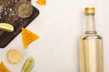 Beyaz mermer yüzeyde ahşap kesim tahtasının yanında limon, tuz ve nachos ile altın tekila manzarası