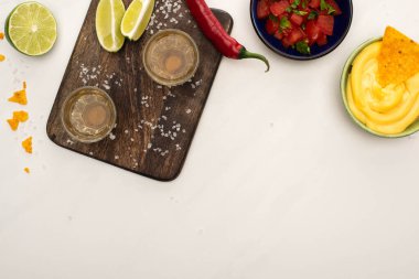 top view of golden tequila with lime, chili pepper, salt and nachos with cheese sauce near wooden cutting board on white marble surface clipart