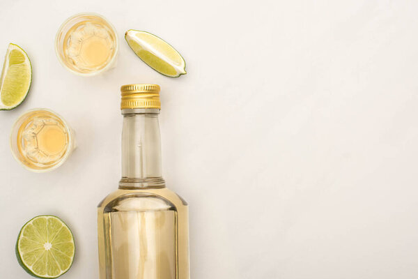 вид сверху на золотую текилу в бутылке и рюмки с лаймом на поверхности белого мрамора
