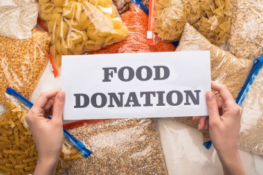 cropped view of woman holding card with food donation lettering near groats and pasta in zipper bags clipart