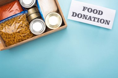 top view of cans and groats in zipper bags in wooden box near card with food donation lettering on blue background clipart