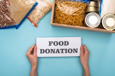 cropped view of woman holding card with food donation lettering near cans and groats in zipper bags in box on blue background clipart