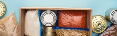 top view of cans and groats in zipper bags in wooden box on blue background, food donation concept clipart