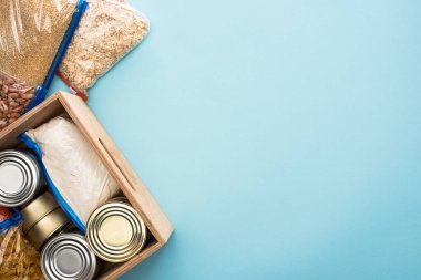 top view of cans and groats in zipper bags in wooden box on blue background, food donation concept clipart