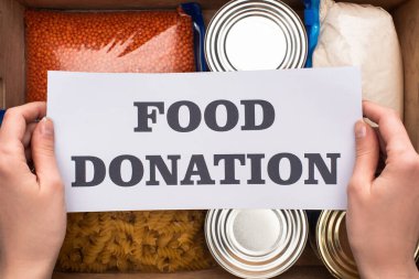 cropped view of woman holding card with food donation lettering above box with cans and groats in zipper bags clipart