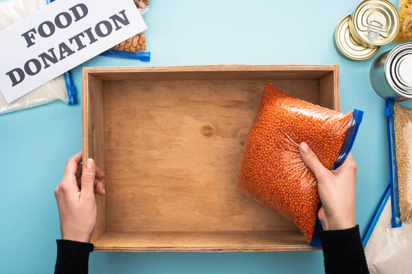 cropped view of woman putting groat in wooden box near card with food donation lettering on blue background