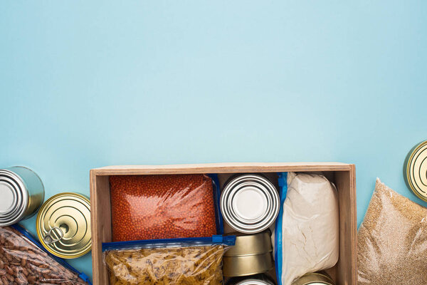 top view of cans and groats in zipper bags in wooden box on blue background, food donation concept