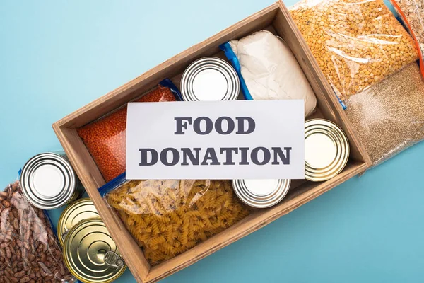 top view of cans and groats in zipper bags in wooden box near card with food donation lettering on blue background