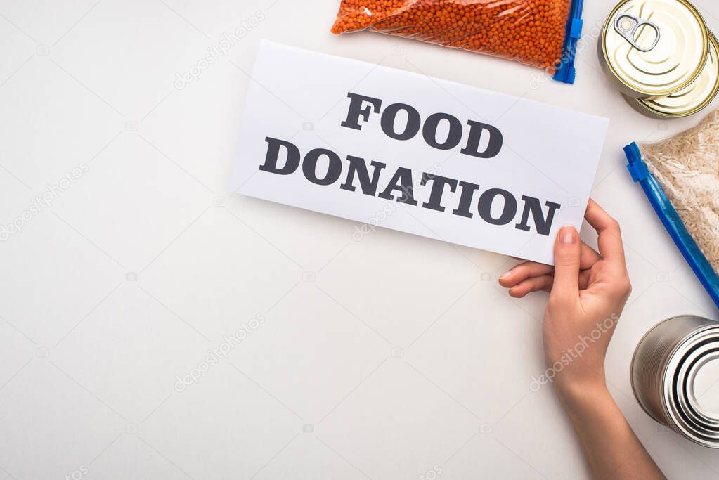 cropped view of woman holding card with food donation lettering near cans and groats in zipper bags on white background