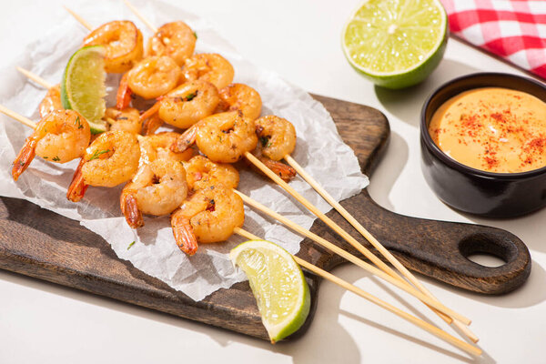 prawns on skewers with lime and sauce on parchment paper on wooden board on white background