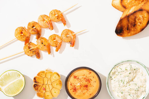 top view of delicious fried prawns on skewers with grilled bread, garlic, lime and sauces on white background