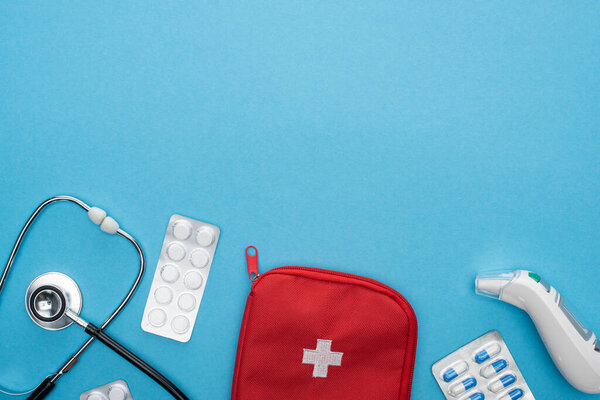 top view of pills in blister packs, stethoscope, first aid kit and ear thermometer on blue background