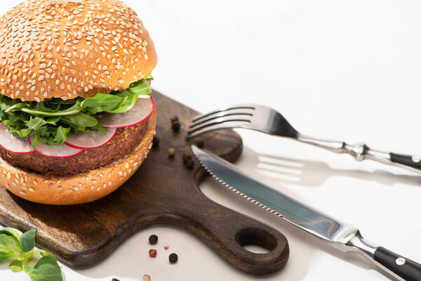 selective focus of delicious vegan burger with radish and arugula on wooden board with black pepper near fork and knife on white background