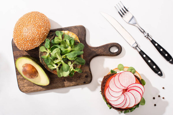 top view of vegan burgers with microgreens, avocado, radish on wooden cutting board on white background with cutlery