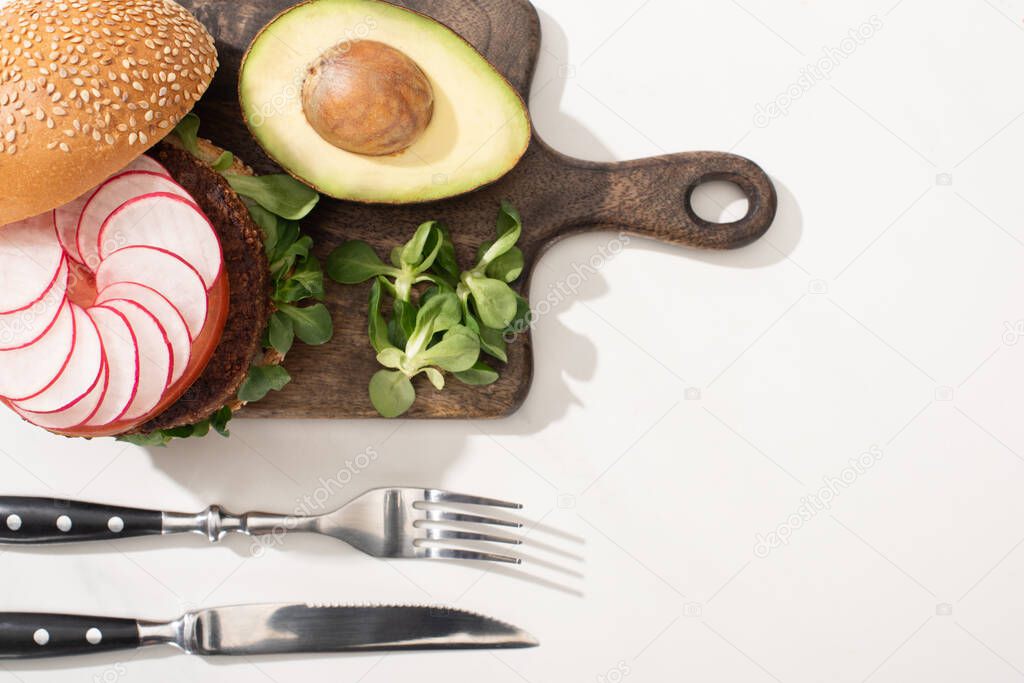 top view of delicious vegan burger with radish, avocado and greens on wooden cutting board near cutlery on white background