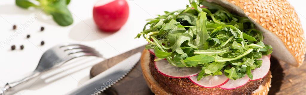 selective focus of delicious vegan burger with radish and arugula on wooden board with black pepper near fork and knife on white background, panoramic shot