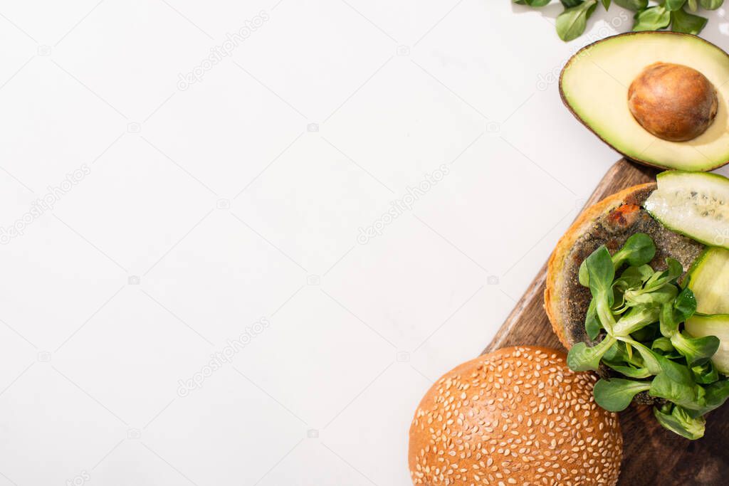 top view of vegan burger with microgreens, cucumber and avocado on wooden cutting board on white background