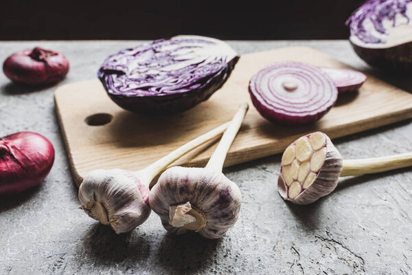 selective focus of red cabbage, onion and garlic on wooden cutting board on grey concrete surface isolated on black