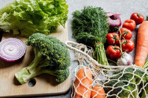fresh ripe vegetables in string bag near cutting board on grey concrete surface