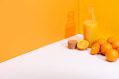olgun portakalların yanında cam şişede taze portakal suyu ve turuncu arka planda beyaz yüzey üzerinde mantar.