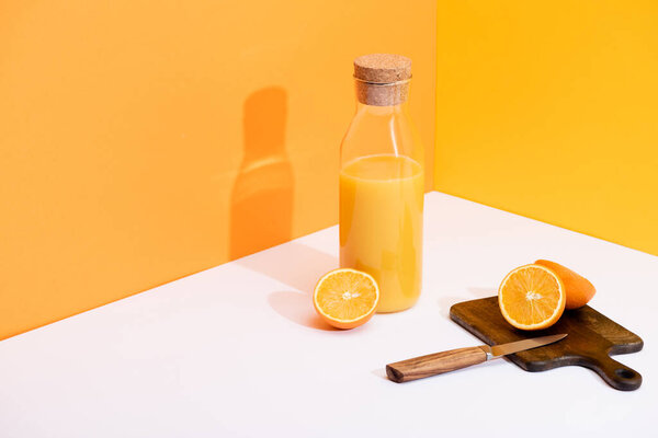 свежий апельсиновый сок в стеклянной бутылке возле спелых апельсинов, деревянная доска с ножом на белой поверхности на оранжевом фоне
