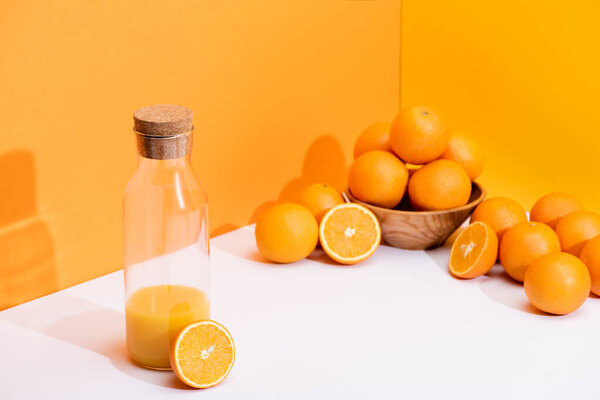 fresh orange juice in glass bottle near ripe oranges in bowl on white surface on orange background
