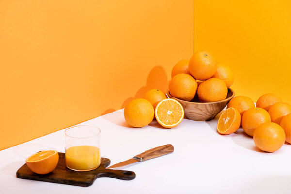 fresh orange juice in glass on wooden cutting board with knife near ripe oranges in bowl on white surface on orange background
