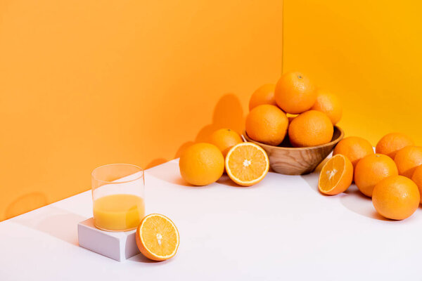 fresh orange juice in glass near ripe oranges in bowl on white surface on orange background