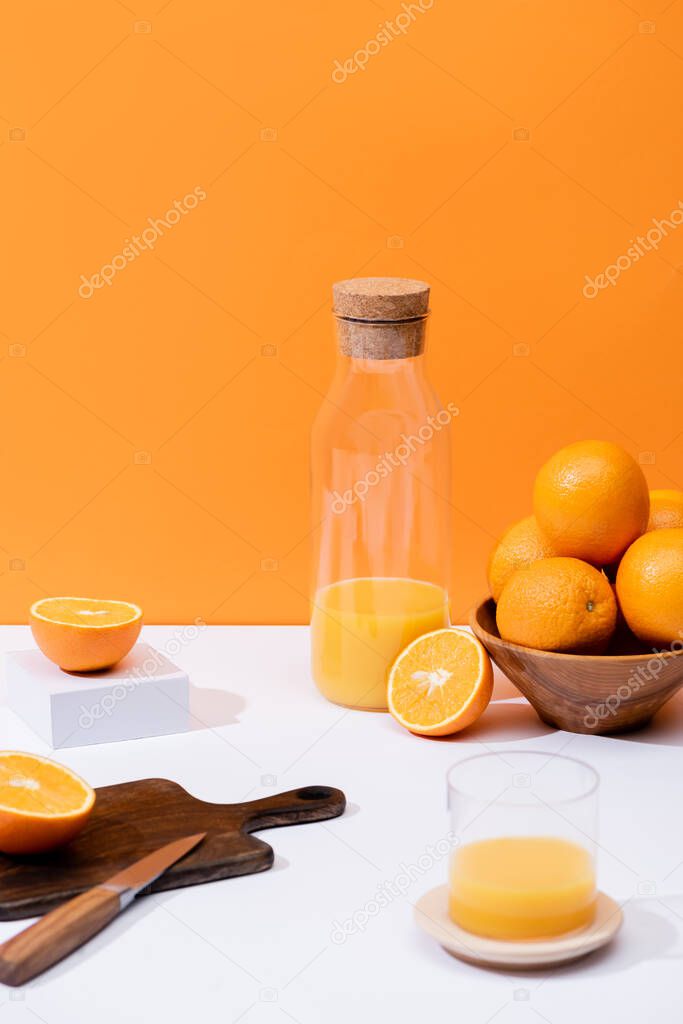fresh orange juice in glass and bottle near oranges in bowl, wooden cutting board with knife on white surface isolated on orange