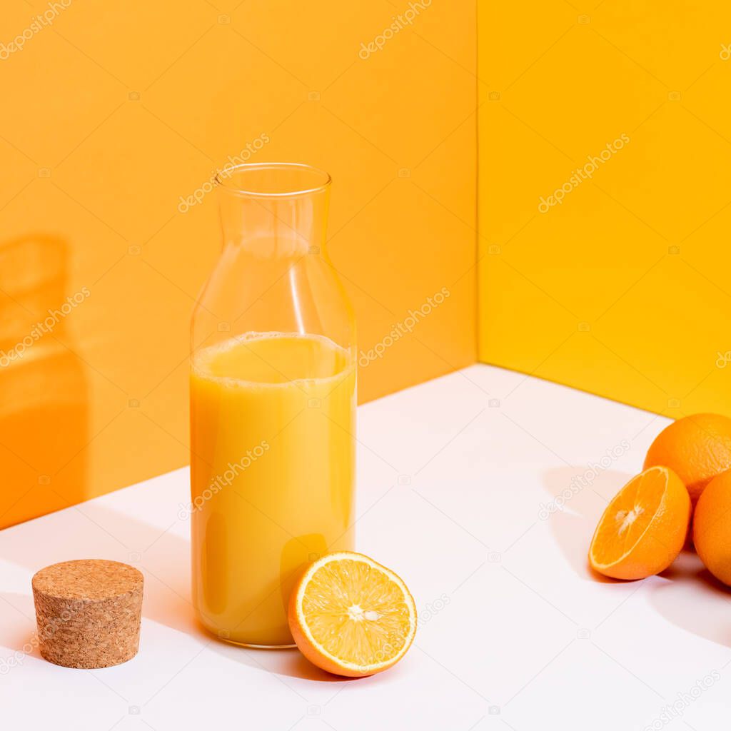 fresh orange juice in glass bottle near ripe oranges and cork on white surface on orange background