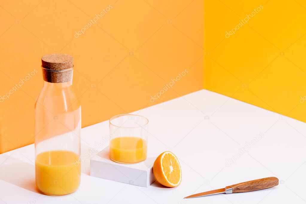 fresh orange juice in glass and bottle near ripe orange and knife on white surface on orange background