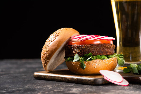 selective focus of tasty vegan burger with vegetables served on wooden cutting board near beer isolated on black