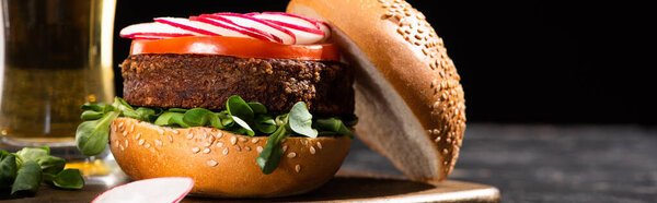 selective focus of tasty vegan burger with vegetables served on wooden cutting board near beer isolated on black, panoramic crop