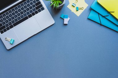 Mavi yüzeydeki fabrika ve ofis malzemelerinin yanındaki dizüstü bilgisayarın üst görüntüsü